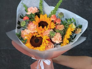 Una explosión de color y sofisticación, donde los girasoles, ranúnculos y claveles se combinan con scabiosas y rúmex en una envoltura blanca y naranja que resalta cada detalle. Este bouquet de Madame Butterfly representa alegría, lujo y creatividad.