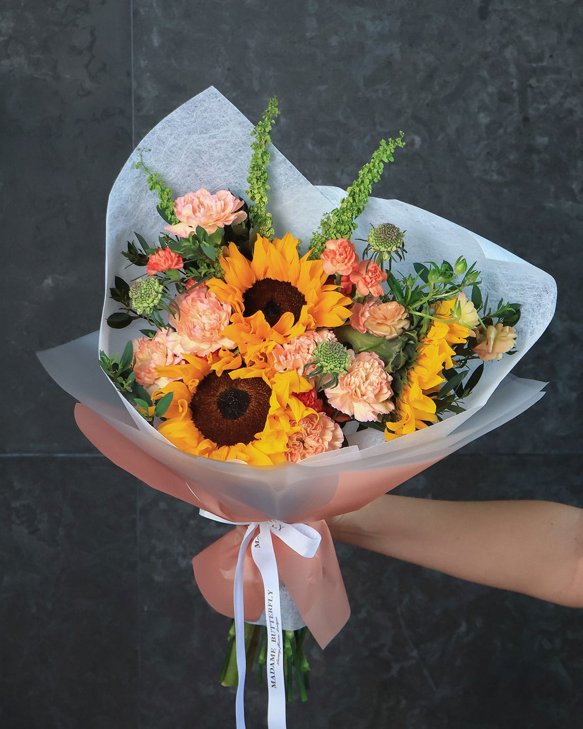 Una explosión de color y sofisticación, donde los girasoles, ranúnculos y claveles se combinan con scabiosas y rúmex en una envoltura blanca y naranja que resalta cada detalle. Este bouquet de Madame Butterfly representa alegría, lujo y creatividad.