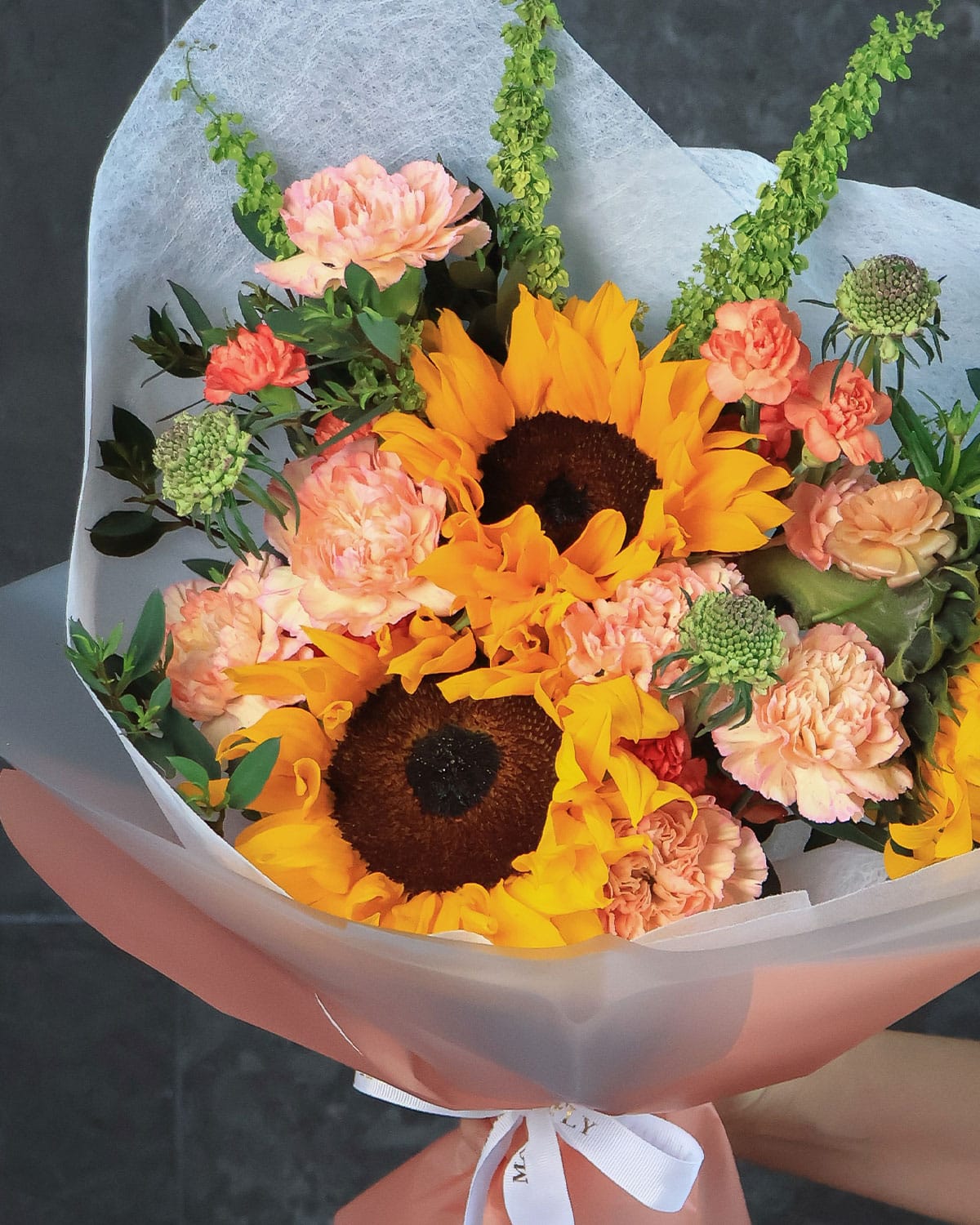 Una explosión de color y sofisticación, donde los girasoles, ranúnculos y claveles se combinan con scabiosas y rúmex en una envoltura blanca y naranja que resalta cada detalle. Este bouquet de Madame Butterfly representa alegría, lujo y creatividad.