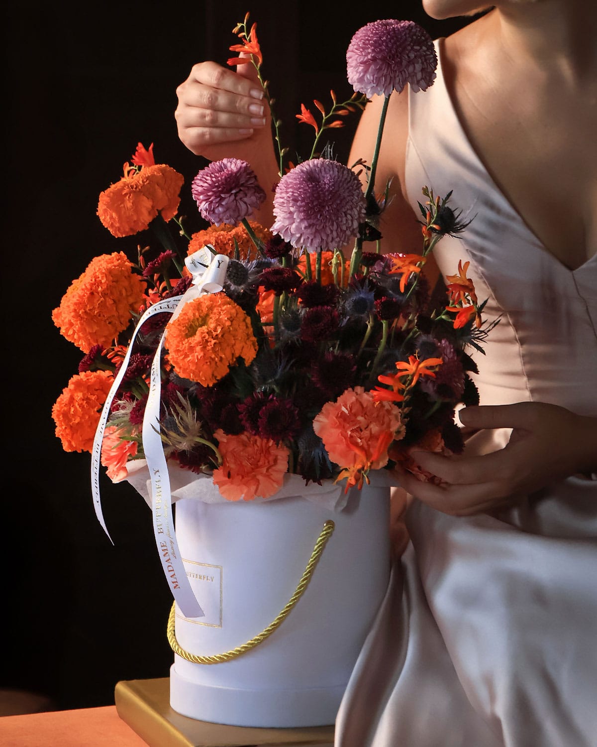 Marigold Solaire de Madame Butterfly es un arreglo de marigold y dhalias en tonos dorados, lilas y morados, sobre una base blanca que exalta su luz. Con crocomia y mini pompones morados, este diseño mezcla lo exótico con lo elegante, creando una pieza audaz, sofisticada y absolutamente inolvidable. Es un homenaje al color, al carácter y a la belleza que no teme brillar.
