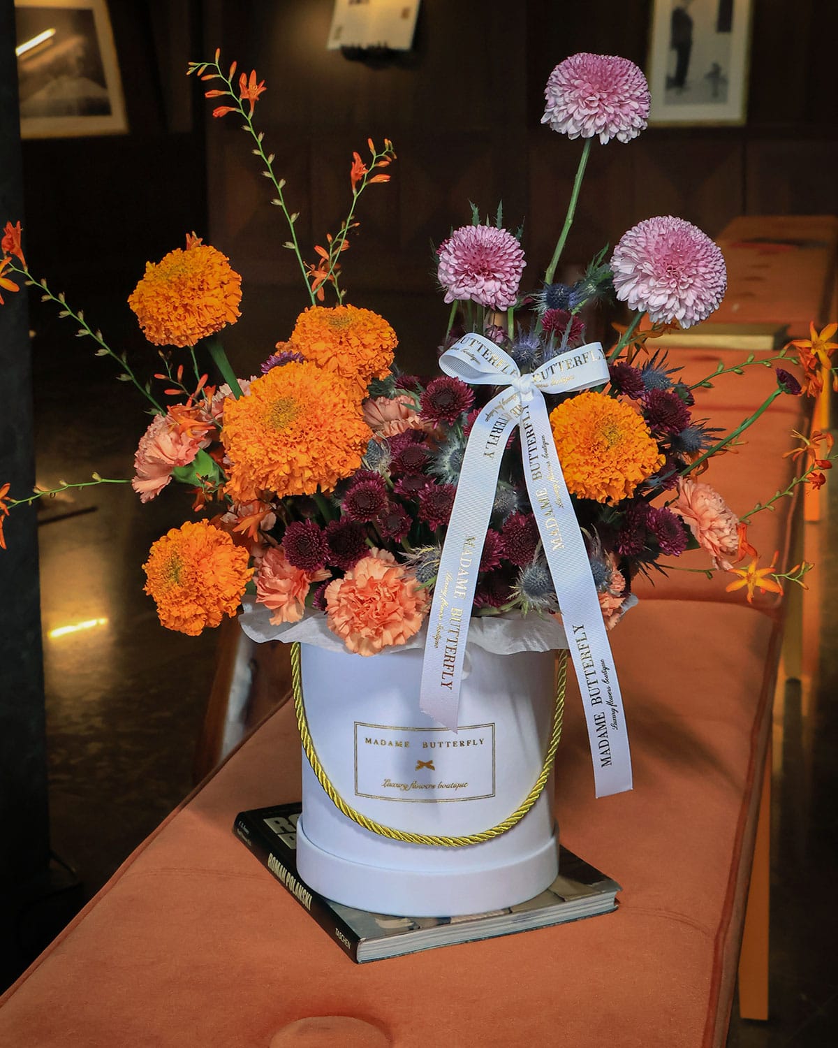 Marigold Solaire de Madame Butterfly es un arreglo de marigold y dhalias en tonos dorados, lilas y morados, sobre una base blanca que exalta su luz. Con crocomia y mini pompones morados, este diseño mezcla lo exótico con lo elegante, creando una pieza audaz, sofisticada y absolutamente inolvidable. Es un homenaje al color, al carácter y a la belleza que no teme brillar.